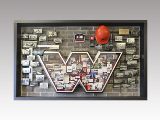 Shadow box collage commemorating Wilhelm Construction's 100th anniversary featuring a silver metal letter "W" on brick with rebar extensions and hard hat