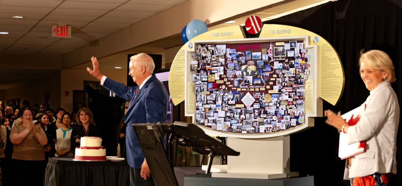 Bill Marriott waving hand and standing next to large shadow box collage on stand for Marriott's 85th Anniversary